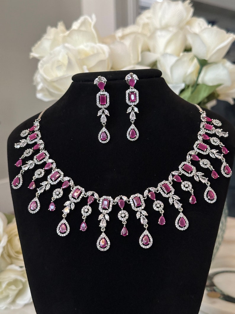 May include: A silver necklace and earrings set with pink cubic zirconia stones. The necklace features a delicate chain with a pendant design and the earrings are drop style.