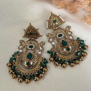 May include: A pair of gold-tone earrings with intricate designs and green and white beads. The earrings feature a crescent moon shape with a large green stone in the center.