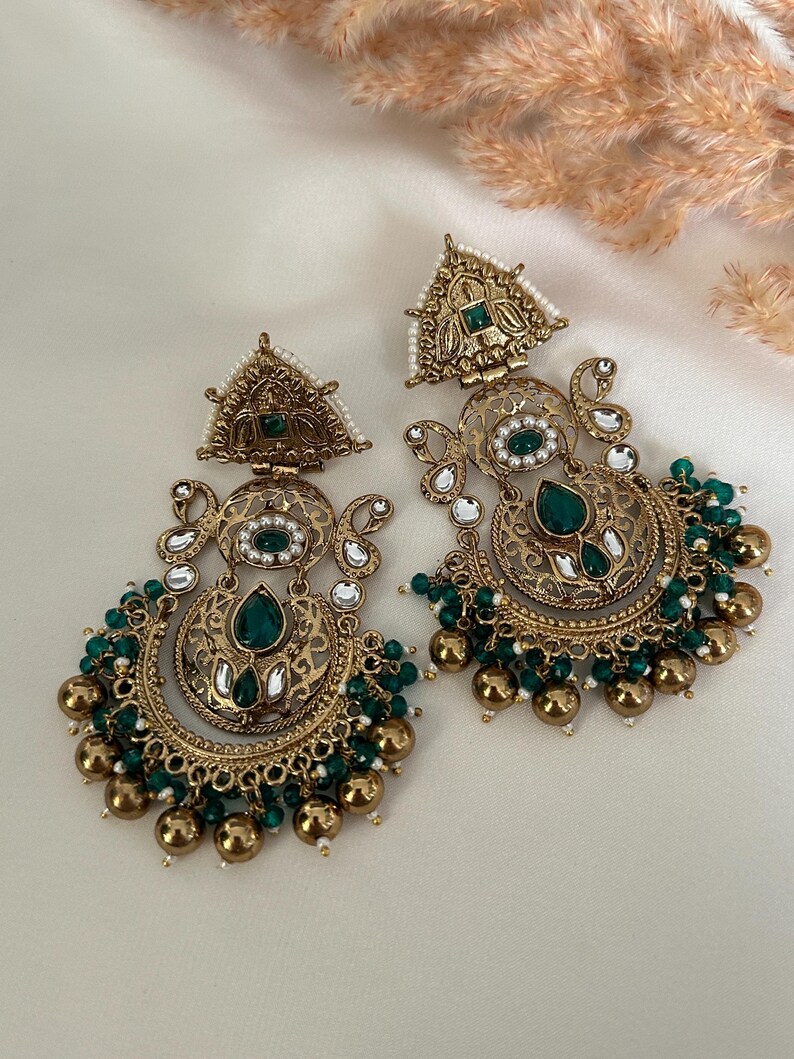 May include: A pair of gold-tone earrings with intricate designs and green and white beads. The earrings feature a crescent moon shape with a large green stone in the center.