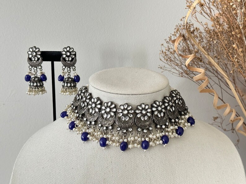 May include: A silver and white choker necklace with blue beads and matching earrings. The necklace features a floral design with pearl accents.