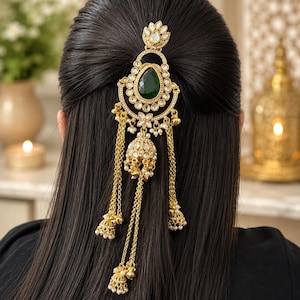 Peut inclure: Un accessoire de cheveux doré orné d'une pierre centrale vert foncé en forme de goutte, entourée de cristaux clairs. Il se compose d'une monture décorative avec des chaînes dorées pendantes et de petites breloques en forme de cloche.