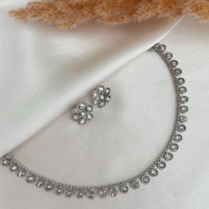 May include: A silver-coloured jewellery set comprising a necklace and matching earrings. The necklace and earrings are embellished with clear, round gemstones. The set is presented on white, silky fabric, with dried ornamental grass in the background.