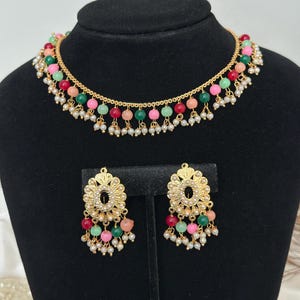 May include: A gold necklace and earrings set with a multi-colored beaded design. The necklace has a delicate chain with a row of pink, green, and red beads. The earrings have a similar design with a gold floral accent.