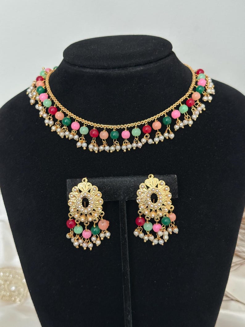 May include: A gold necklace and earrings set with a multi-colored beaded design. The necklace has a delicate chain with a row of pink, green, and red beads. The earrings have a similar design with a gold floral accent.