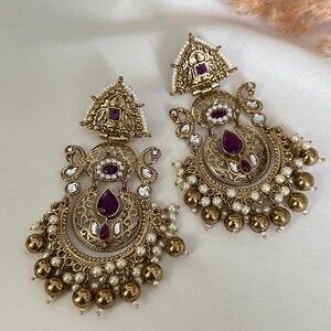 May include: A pair of gold-tone earrings with intricate filigree designs. The earrings feature a crescent moon shape with a purple gemstone at the center. The earrings are adorned with small white pearls and gold beads.