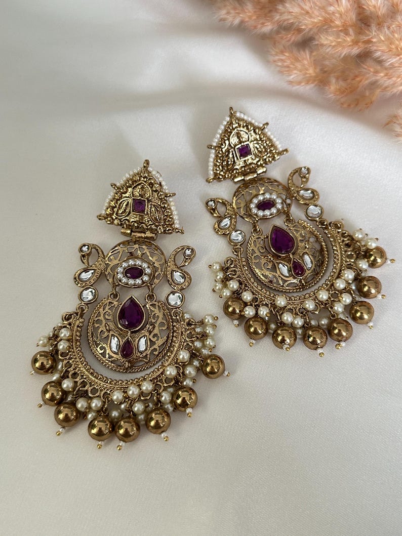 May include: A pair of gold-tone earrings with intricate filigree designs. The earrings feature a crescent moon shape with a purple gemstone at the center. The earrings are adorned with small white pearls and gold beads.