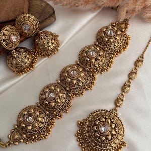 May include: A gold-coloured jewellery set comprising a necklace, earrings, and a headpiece. The set features detailed floral and beaded patterns, with clear stone accents. The earrings are round with a dangling bell shape. The necklace is made up of five large, round elements.