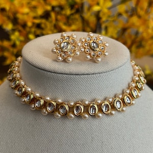 May include: A gold and white pearl choker necklace with a decorative design of round, clear stones. The necklace is displayed on a white mannequin bust. Matching earrings are also shown.
