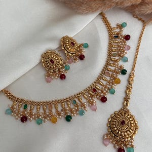 Può includere: Un set di gioielli dorati composto da collana, orecchini e copricapo. Il set è adornato con perline colorate nei toni del rosso, verde, giallo e rosa, insieme a piccole perle bianche. I gioielli sono esposti su una superficie di tessuto bianco.