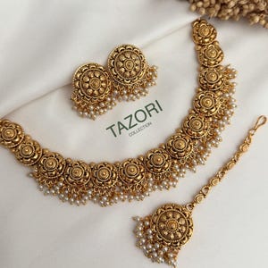 Könnte beinhalten: Ein goldfarbenes Schmuckset mit Halskette, Ohrringen und einem Kopfschmuck. Das Set hat ein Blumenmuster mit kleinen weißen Perlenakzenten. Die Ohrringe sind rund, und die Halskette und der Kopfschmuck haben ein ähnliches Design. Der Text "TAZORI COLLECTION" ist sichtbar.
