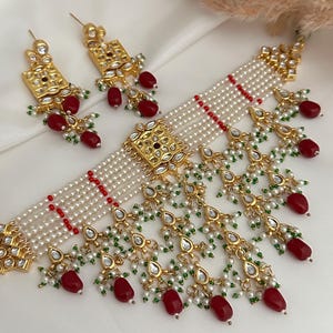 Puede incluir: Un collar gargantilla de perlas blancas y doradas con detalles rojos y verdes. El collar tiene un gran colgante adornado en el centro y está decorado con pequeñas cuentas colgantes.