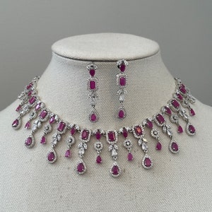 May include: A silver necklace and earrings set with a dangling design. The necklace features a row of square-shaped red gemstones with smaller red gemstones and clear stones hanging from the bottom. The earrings have a similar design with a single square-shaped red gemstone and smaller red gemstones and clear stones hanging from the bottom.