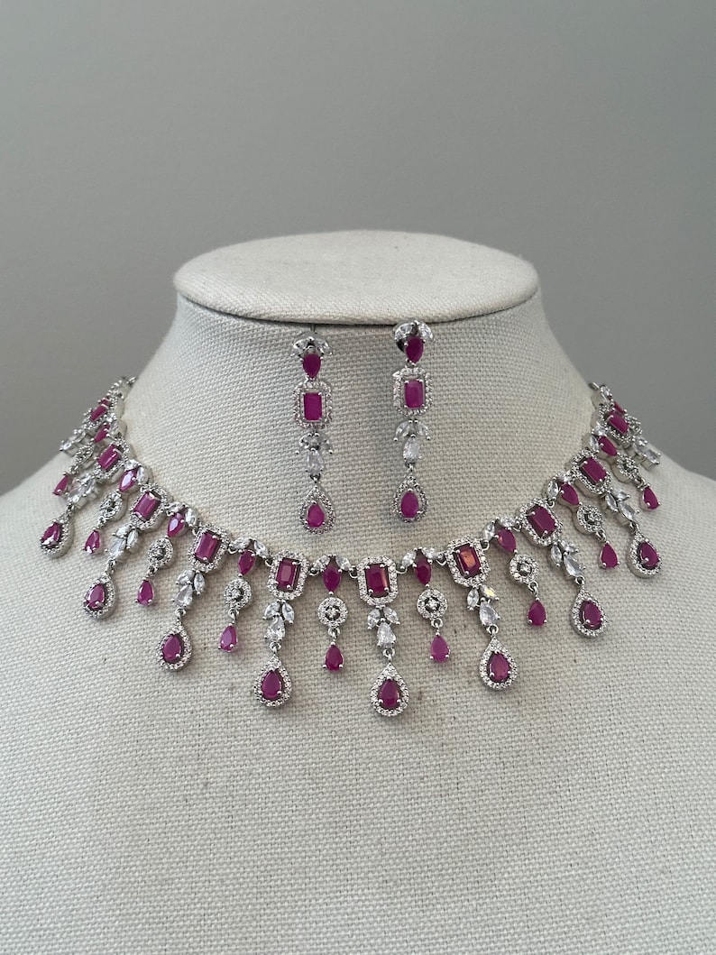 May include: A silver necklace and earrings set with a dangling design. The necklace features a row of square-shaped red gemstones with smaller red gemstones and clear stones hanging from the bottom. The earrings have a similar design with a single square-shaped red gemstone and smaller red gemstones and clear stones hanging from the bottom.