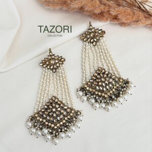 May include: A pair of long, ornate earrings featuring a diamond-shaped top and bottom section with antique gold-tone metal, embedded with clear crystals. Multiple strands of small white pearls connect the sections, with larger pearls dangling below.