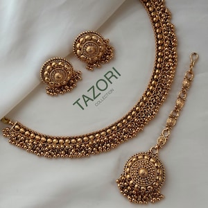 May include: A gold-toned jewelry set featuring a necklace, earrings, and a headpiece. The set includes a necklace with a beaded design, matching stud earrings, and a headpiece with a central pendant. The jewelry is displayed on a white surface, with the text "TAZORI COLLECTION".