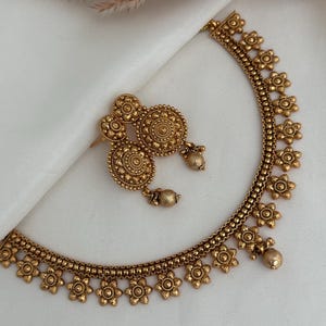 Peut inclure: Un ensemble de bijoux doré comprenant un collier et des boucles d'oreilles assorties. Le collier et les boucles d'oreilles présentent un motif floral avec des détails complexes. L'ensemble est présenté sur une surface blanche, mettant en valeur la couleur métallique chaude et le design.