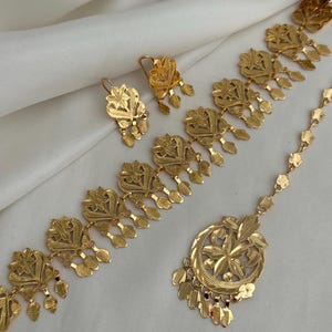 May include: A gold-toned jewelry set consisting of a necklace, a pair of drop earrings, and a maang tikka. Each piece features repeating floral-inspired motifs with dangling leaf-shaped charms, all crafted with a textured, metallic finish.