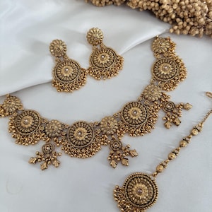 May include: A gold-toned jewelry set featuring a necklace, earrings, and a headpiece. The necklace and earrings have a floral design with dangling accents. The headpiece has a central floral element. The set is displayed on a white surface.