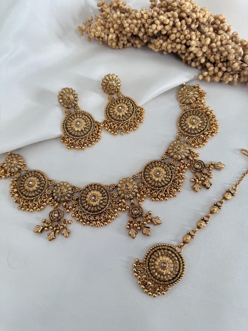 May include: A gold-toned jewelry set featuring a necklace, earrings, and a headpiece. The necklace and earrings have a floral design with dangling accents. The headpiece has a central floral element. The set is displayed on a white surface.