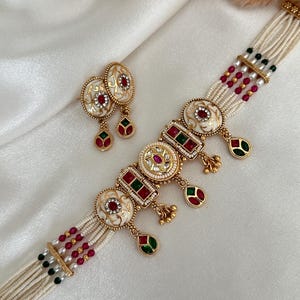 Puede incluir: Un collar gargantilla blanco y dorado con piedras rojas y verdes. El collar tiene un diseño de múltiples hilos con perlas blancas y cuentas rojas y verdes. El colgante presenta un diseño de esmalte blanco con piedras rojas y verdes. También se incluyen pendientes a juego con un diseño de esmalte blanco y piedras rojas y verdes.