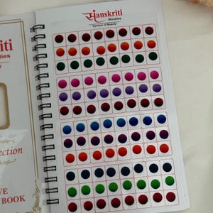 May include: A spiral-bound book of bindis in various colors, including red, orange, green, pink, purple, blue, and black. The brand name "Hanskriti Bindies" is printed at the top. The book is open, displaying multiple rows of small, round bindis.