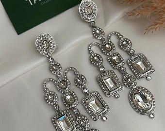 May include: A pair of silver chandelier earrings with clear crystal stones. The earrings are made of metal and have a dangling design. The earrings are from the Tazori Collection.