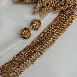 Könnte beinhalten: Goldfarbenes Schmuckset mit Halskette und passenden Ohrringen. Die Halskette hat ein detailliertes Blumendesign mit hängenden Akzenten. Die Ohrringe sind rund, mit einem ähnlichen Blumenmuster. Das Set wird auf einer weißen Oberfläche präsentiert.