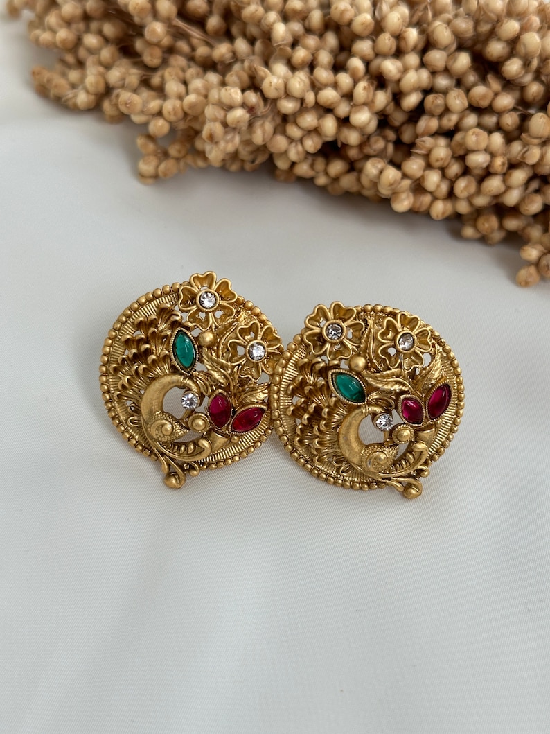 Puede incluir: Un par de pendientes de tono dorado con un dise&ntilde;o de pavo real. Los pendientes est&aacute;n adornados con piedras preciosas verdes y rojas y diamantes de imitaci&oacute;n blancos.