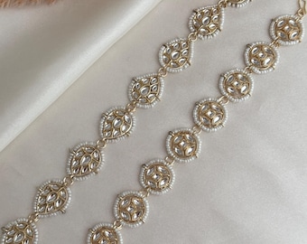 May include: Two gold headbands with clear, reflective details and small white beads. The headbands have a repeating pattern of teardrop and circular shapes. The jewellery is displayed on a white, silky fabric.
