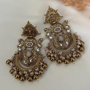 May include: A pair of gold-tone earrings with a pink and white design. The earrings feature a crescent moon shape with intricate details and a dangling chain of beads.