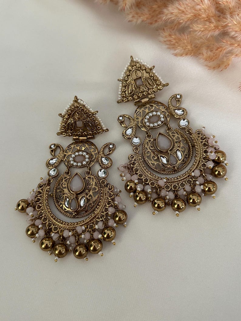 May include: A pair of gold-tone earrings with a pink and white design. The earrings feature a crescent moon shape with intricate details and a dangling chain of beads.