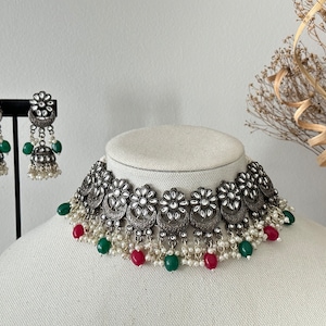 May include: A silver choker necklace with a floral design and pearl accents. The necklace has green and red beads hanging from it. Matching earrings with a similar design are also included.