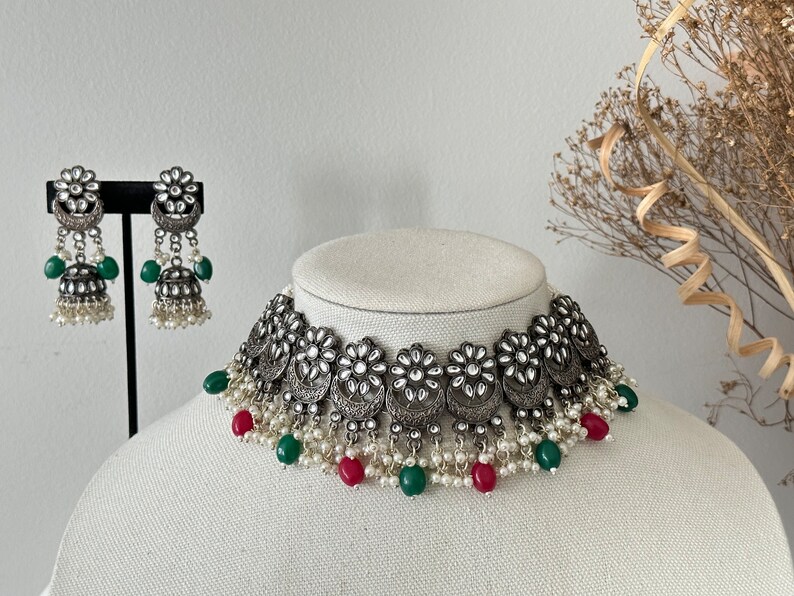 May include: A silver choker necklace with a floral design and pearl accents. The necklace has green and red beads hanging from it. Matching earrings with a similar design are also included.