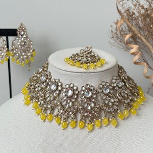 May include: A gold and white necklace with yellow beads, matching earrings, and a matching maang tikka. The necklace is made of metal and has a floral design. The earrings are dangle earrings and the maang tikka is a small piece of jewelry that is worn on the forehead.