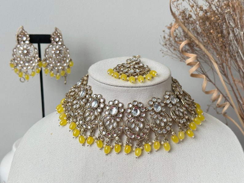 May include: A gold and white necklace with yellow beads, matching earrings, and a matching maang tikka. The necklace is made of metal and has a floral design. The earrings are dangle earrings and the maang tikka is a small piece of jewelry that is worn on the forehead.