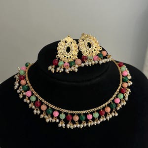May include: A gold necklace with a delicate chain and colorful beads. The necklace has a dangling design with small pearls and colorful beads. The necklace comes with a matching pair of earrings with a similar design.