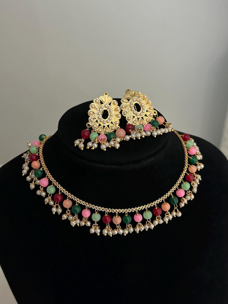 May include: A gold necklace with a delicate chain and colorful beads. The necklace has a dangling design with small pearls and colorful beads. The necklace comes with a matching pair of earrings with a similar design.