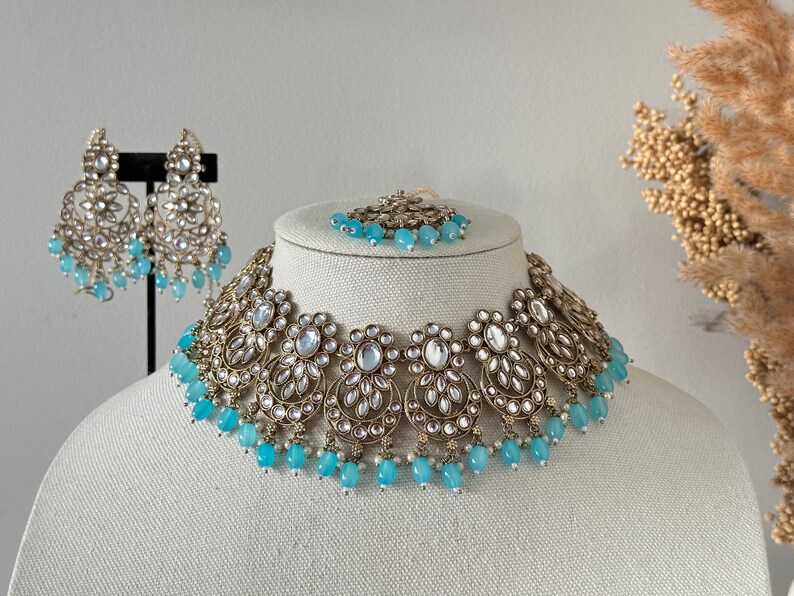 May include: A gold and silver necklace with blue beads and a matching pair of earrings. The necklace is made of intricate metalwork and features a floral design.