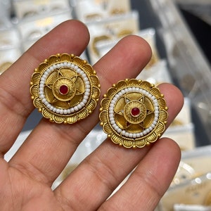 Puede incluir: Un par de pendientes de color dorado con un dise&ntilde;o circular. Cada pendiente presenta una piedra preciosa roja en el centro rodeada de un c&iacute;rculo de cuentas blancas y un borde festoneado decorativo.