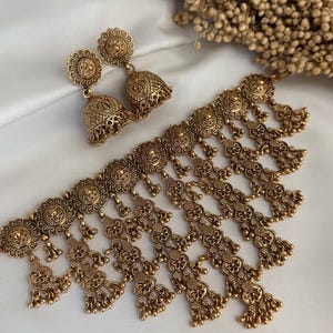 Op de afbeelding: Een goudkleurige metalen sieradenset bestaande uit een choker-halsketting en een paar jhumka-oorbellen. De ketting is voorzien van ronde medaillons met ingewikkelde reliëfpatronen en meerdere bungelende bloemvormige bedels met kleine kraaltjes.