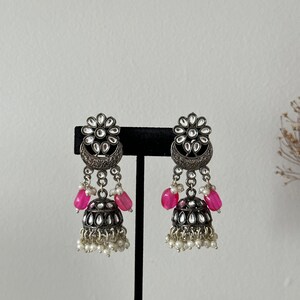 May include: A pair of silver earrings with pink and white beads. The earrings feature a floral design with a crescent moon shape and a dangling bell charm.