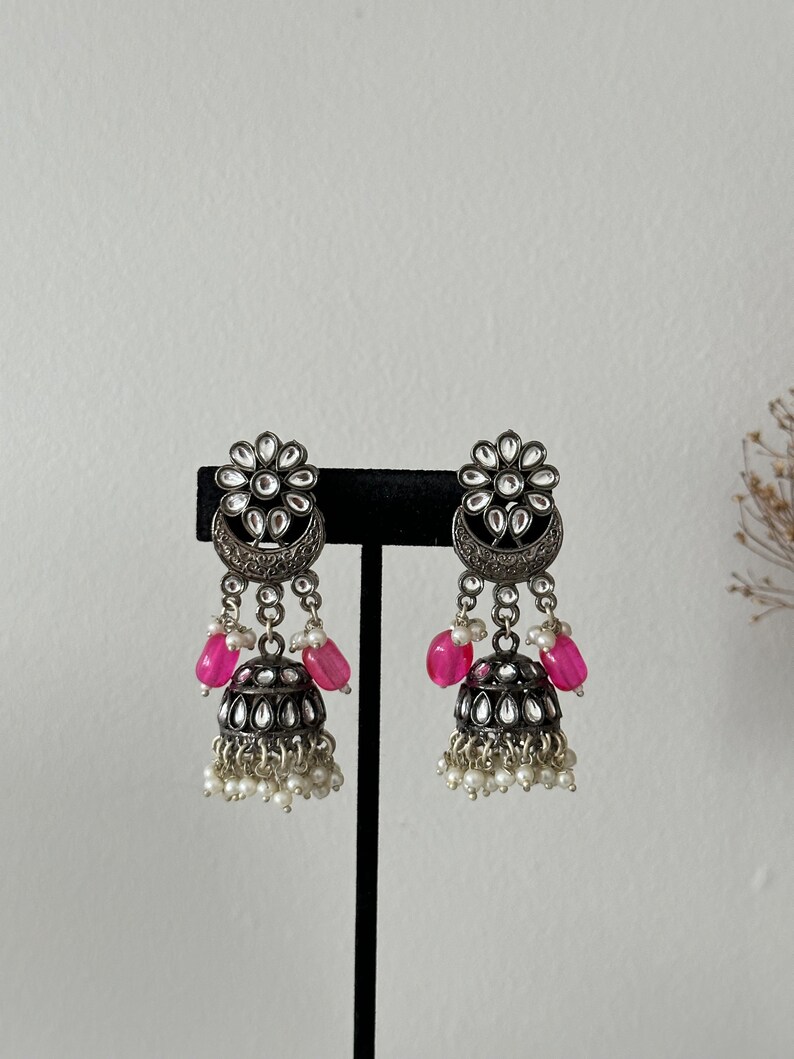 May include: A pair of silver earrings with pink and white beads. The earrings feature a floral design with a crescent moon shape and a dangling bell charm.