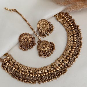 May include: A gold-toned necklace set with matching earrings and a maang tikka. The necklace features a layered design with small round beads and floral motifs. The earrings and maang tikka have a similar design with a round centre piece and dangling beads.