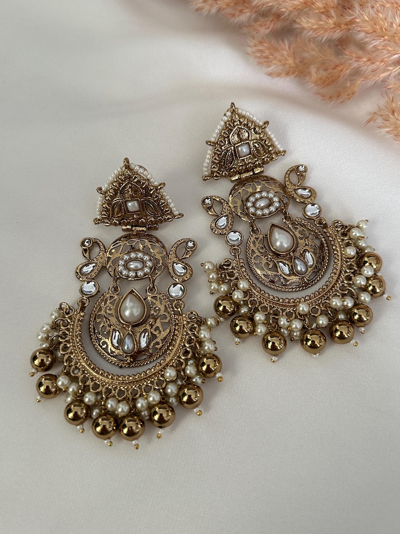 May include: A pair of gold-tone earrings with intricate filigree designs and pearl accents. The earrings feature a crescent moon shape with a teardrop-shaped pearl pendant.
