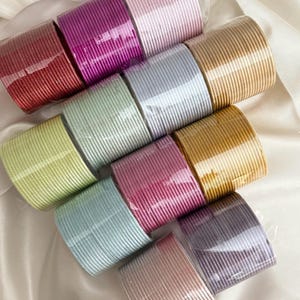 Könnte beinhalten: Eine Sammlung farbenfroher, gerippter Stoffrollen in verschiedenen Farbtönen, darunter Rot, Lila, Rosa und Gold. Die Rollen sind in klarem Kunststoff verpackt und auf einer weißen, satinartigen Oberfläche angeordnet. Der Stoff scheint eine dekorative Zierleiste oder ein Band zu sein.