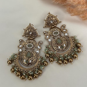May include: A pair of gold-tone earrings with intricate designs and green and white stones. The earrings feature a crescent moon shape with dangling beads.