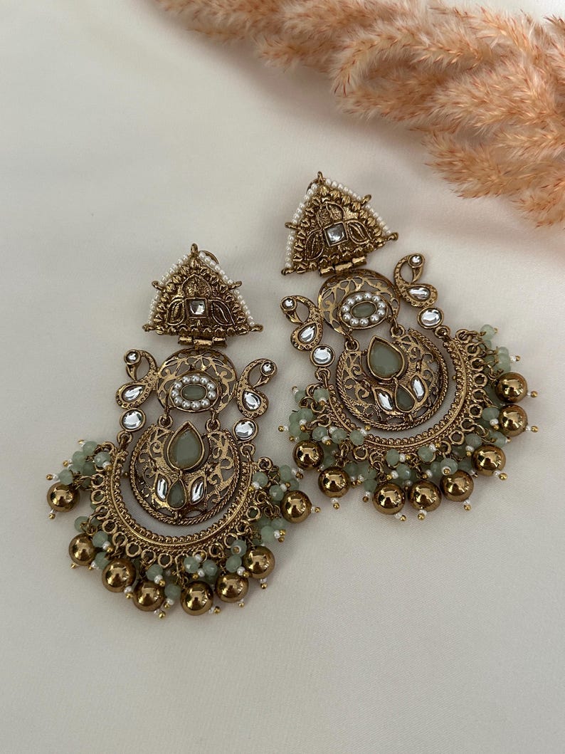 May include: A pair of gold-tone earrings with intricate designs and green and white stones. The earrings feature a crescent moon shape with dangling beads.