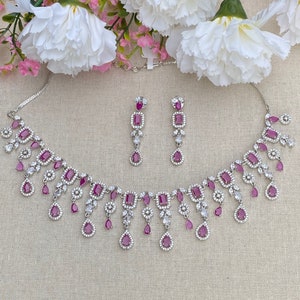 May include: A silver necklace and earrings set with a dangling design. The necklace features a row of square-shaped pink stones surrounded by smaller clear stones. The earrings have a similar design with a single pink stone.