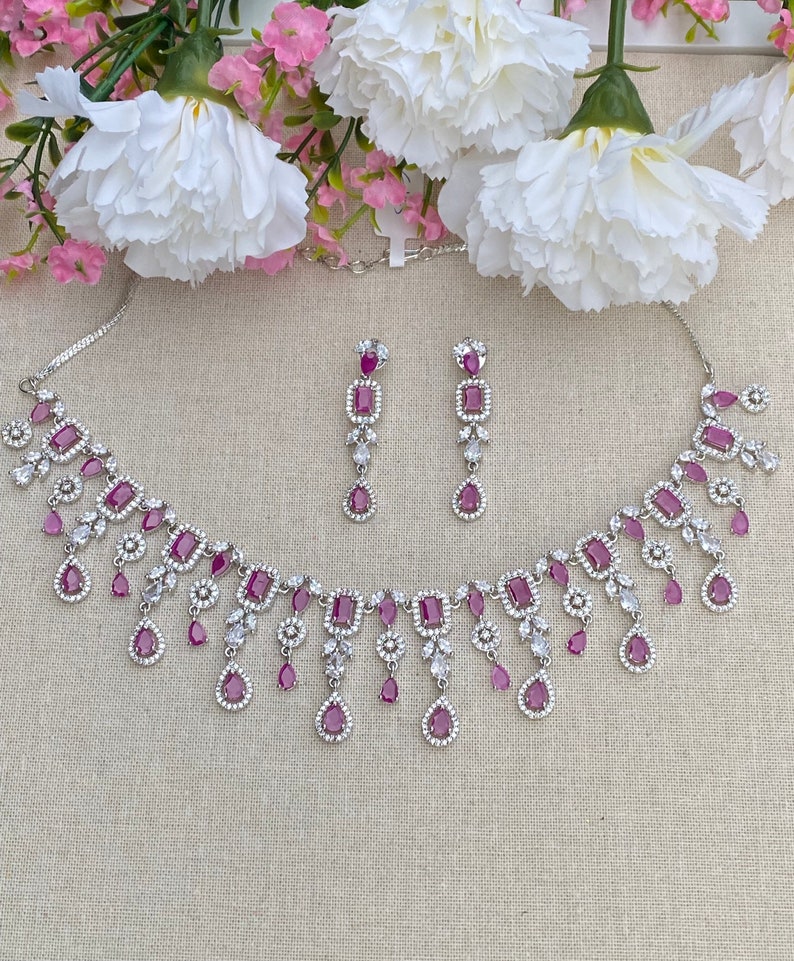 May include: A silver necklace and earrings set with a dangling design. The necklace features a row of square-shaped pink stones surrounded by smaller clear stones. The earrings have a similar design with a single pink stone.