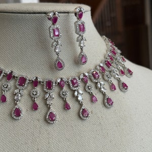 May include: A silver necklace with a row of pink and white stones, and matching earrings with pink stones. The necklace has a dangling design with multiple drops of pink and white stones.